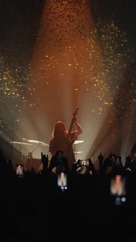 Una banda en el escenario entre luces ámbar y confeti, con el público al frente